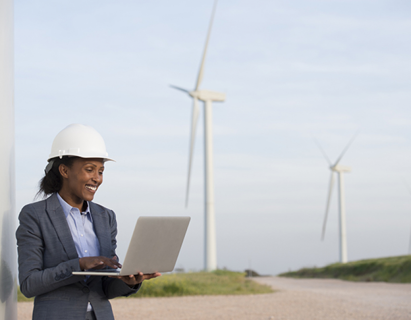 Sustainable energy engineer with laptop PC at a wind farm in a rural landscape. Worker monitoring, checking, analyzing data online from wind-powered turbines.