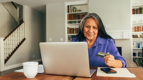 A senior adult woman sits at the dining table paying bills and banking using her mobile laptop. She is using his credit card for his online banking, paying of bills, and shopping.