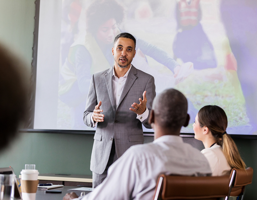 Mid adult businessman discusses a community cleanup event with his colleague. A projected image of people cleaning up the environment is on a screen in the background.