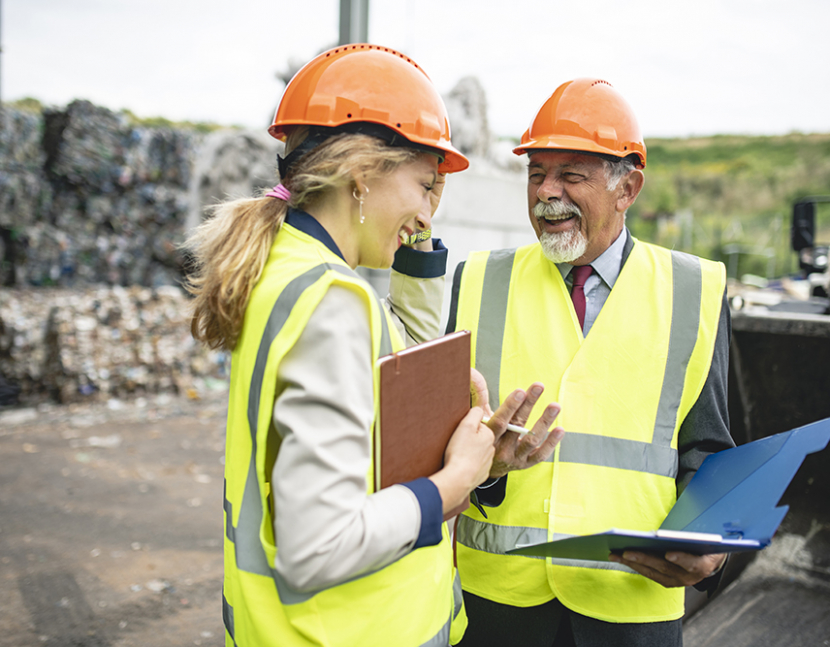 Smiling waste management quality controllers sharing results outdoors with stacks of baled recyclables in background.