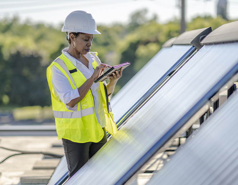 A professional woman wearing a hard hat and a safety vest holds a pen and works on a tablet while surveying solar panels.
