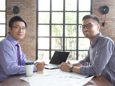 Closeup of two smiling business men looking at camera, discussing issues and working at office desk with brick walls and loft windows in background. Side view.