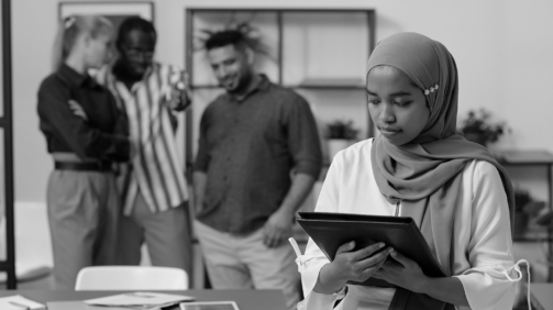 Young Muslim businesswoman in hijab holding black folder with documents against group of intercultural colleagues mocking at her