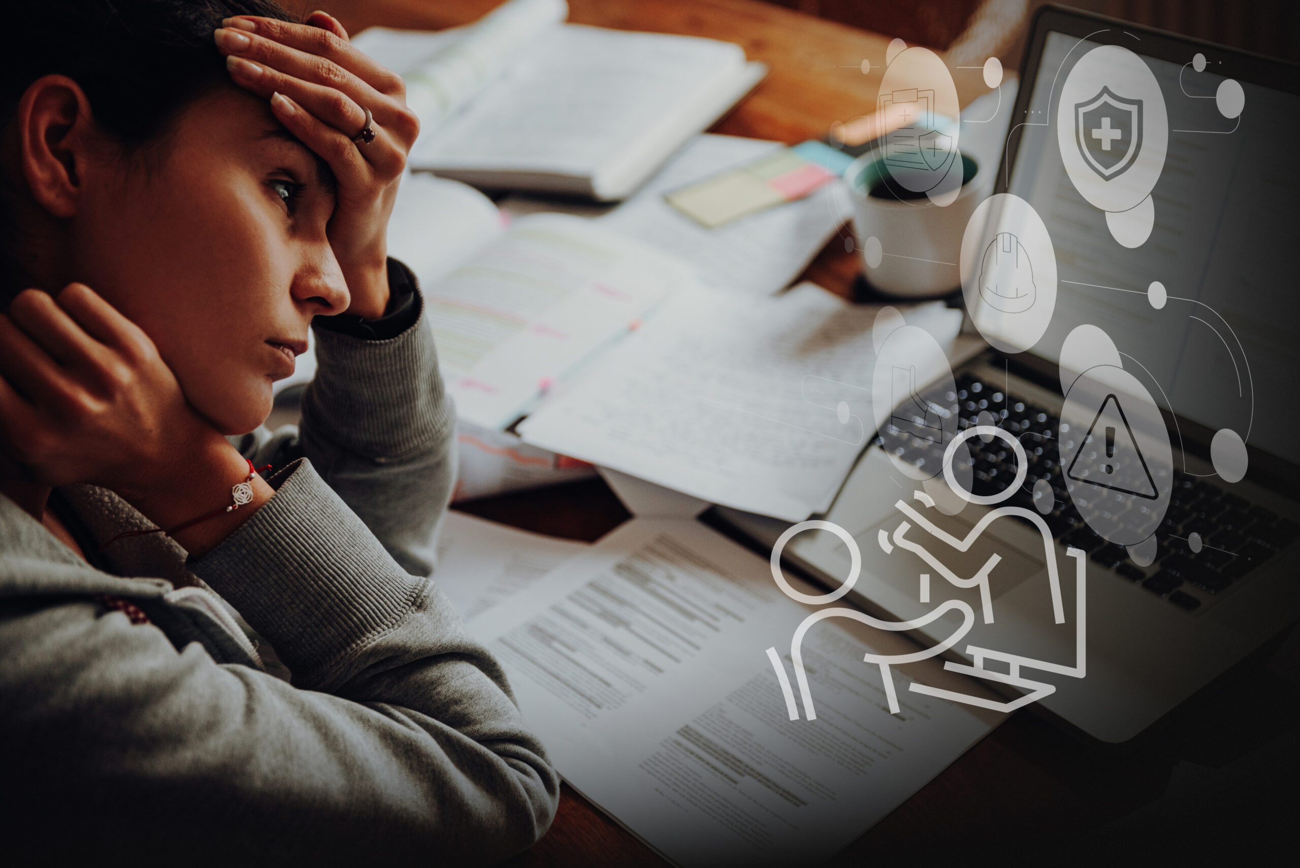 Person sits at a desk with open papers, holding their head in distress while studying. A laptop and notes are on the desk with faint digital icons overlaid on the screen.