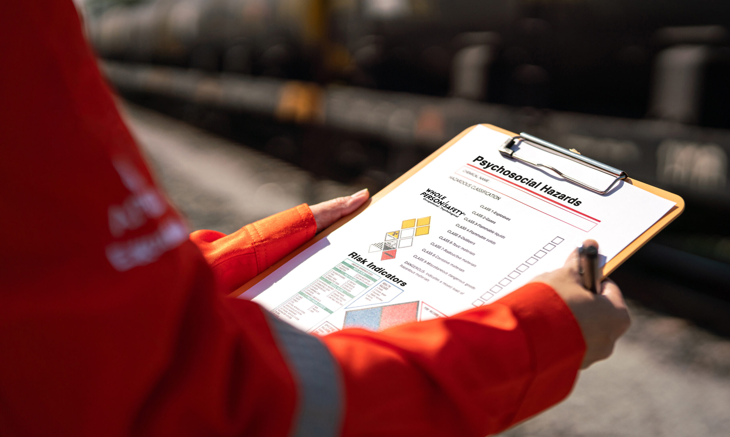 Person in an orange jacket holds a clipboard with a document labeled Psychosocial Hazards and risk icons at the bottom edge.