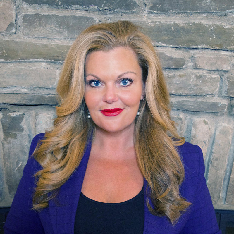 Portrait of a smiling blonde woman with blue eyes and red lipstick, wearing a purple blazer against a stone wall background.