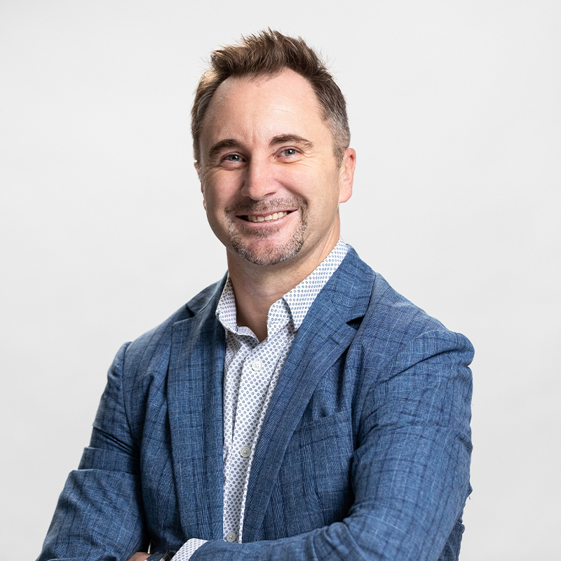 Smiling man in a blue blazer and patterned shirt, posing against a light neutral background.