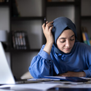 young muslim businesswoman in headscarf working with her computer in home office | TeamsynerG Global Consulting Woman wearing a blue hijab sits at a desk with papers and a laptop, holding a pen and appearing focused or thoughtful. Bookshelves are visible in the background.