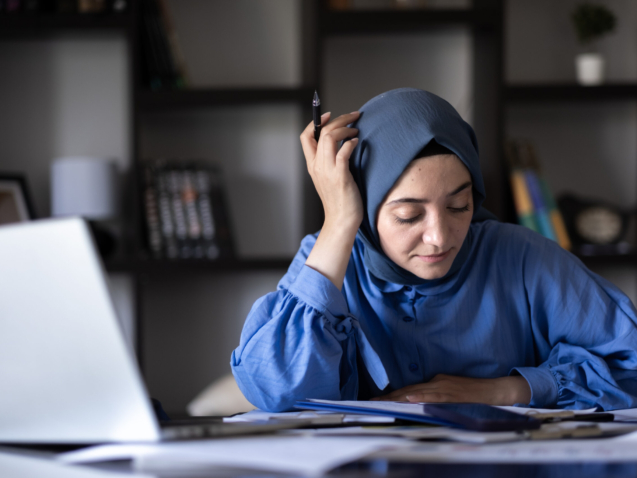 young muslim businesswoman in headscarf working with her computer in home office | TeamsynerG Global Consulting Woman wearing a blue hijab sits at a desk with papers and a laptop, holding a pen and appearing focused or thoughtful. Bookshelves are visible in the background.