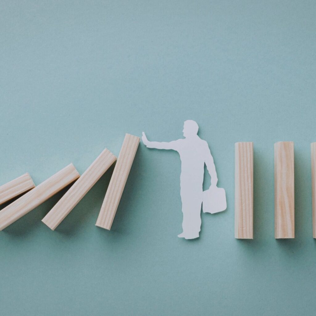 White paper silhouette of a person pushing a domino to stop a falling chain of wooden blocks on a blue background.