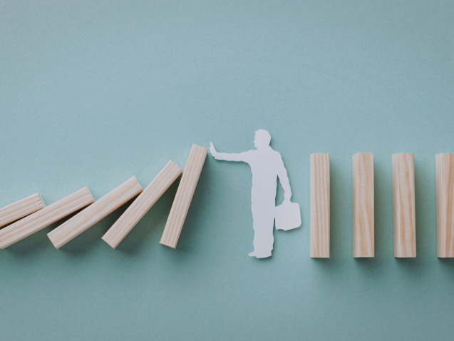 TeamsynerG_Why Your Psychosocial Risk Interventions Keep Starting Over_Banner_No text | TeamsynerG Global Consulting White paper silhouette of a person pushing a domino to stop a falling chain of wooden blocks on a blue background.