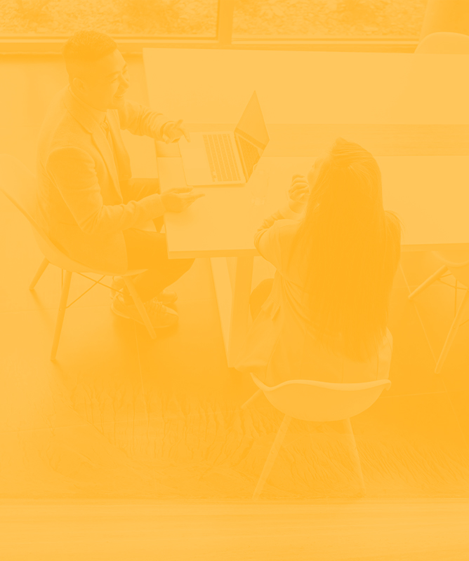 Two professionals sit at a table, reviewing a laptop and a document in a bright office.
