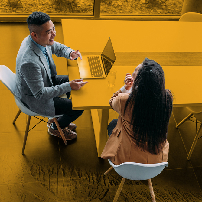 Two colleagues in a yellow-toned office discuss at a table with an open laptop between them.