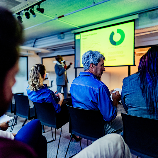People seated in a conference room watching a presenter on stage with a projected slide showing a large green circular chart.