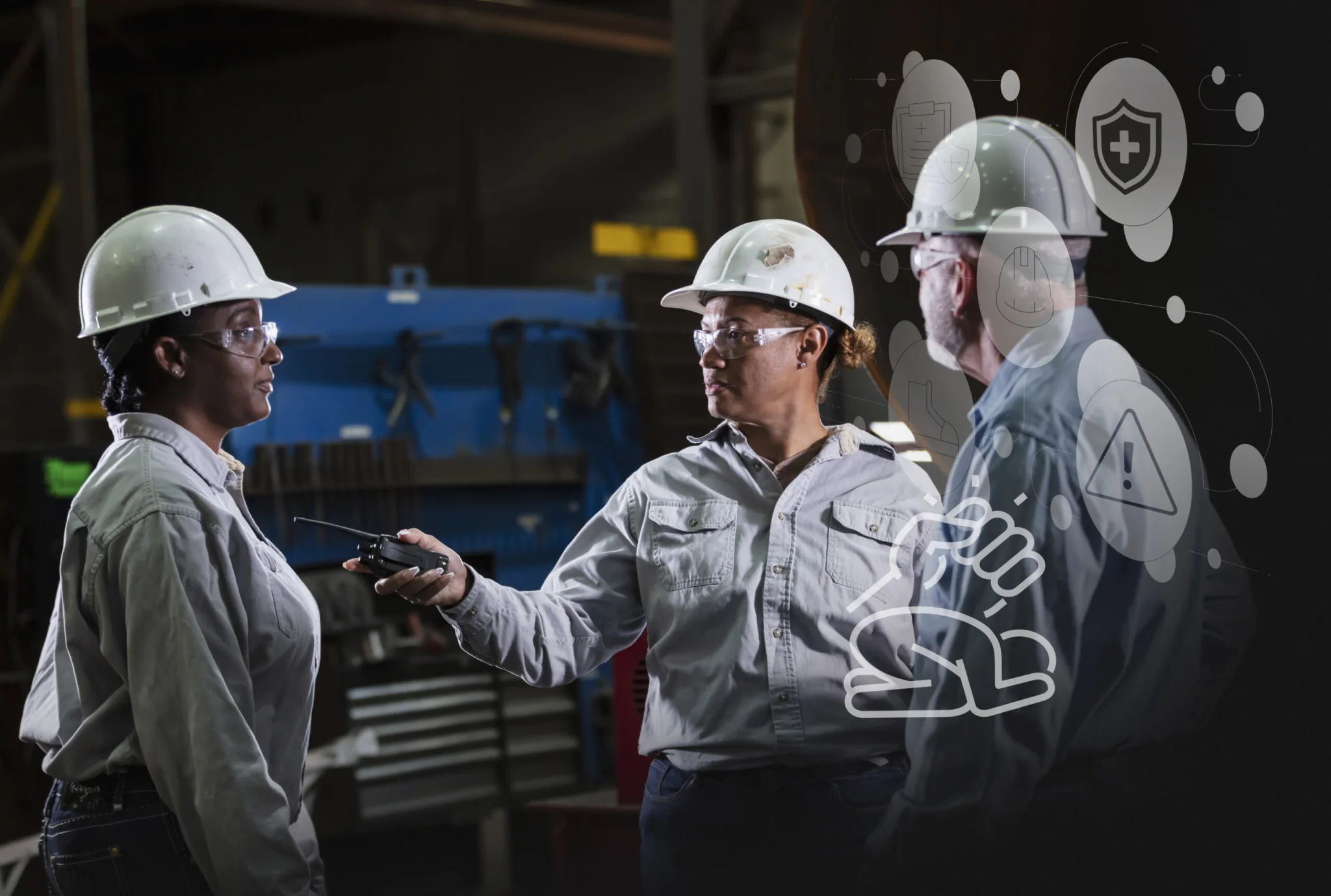 Three factory workers in hard hats and safety glasses converse, one holding a handheld radio in a dim industrial setting with equipment in the background.